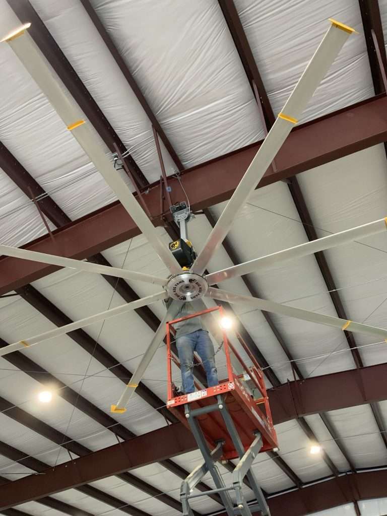 Electrician on a lift installing an HVLS fan beneath steel ceiling trusses