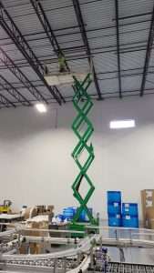 Electrician on a tall scissor lift working on ceiling electrical above a conveyor system