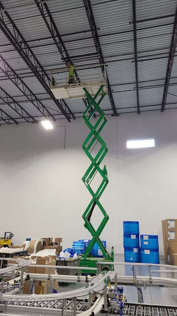Electrician on a tall scissor lift working on ceiling electrical above a conveyor system