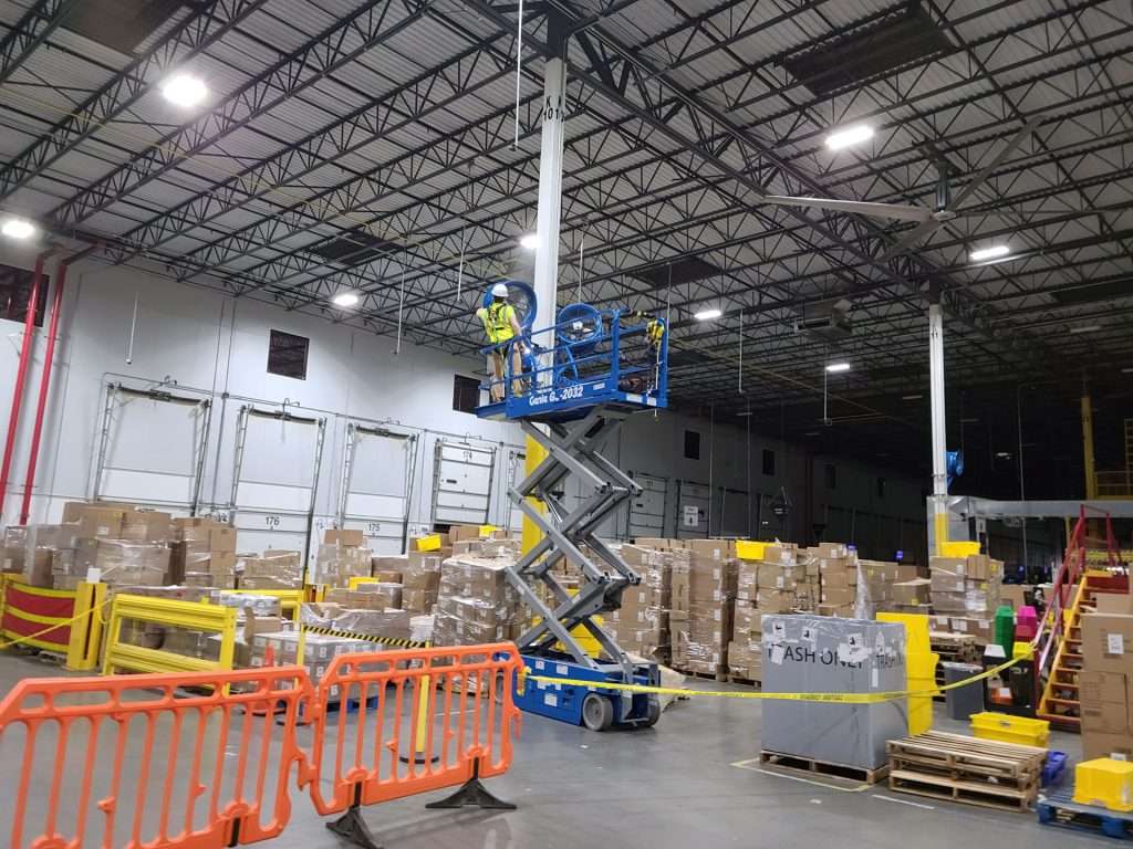 Electrician on a scissor lift performing overhead electrical work in a busy distribution warehouse