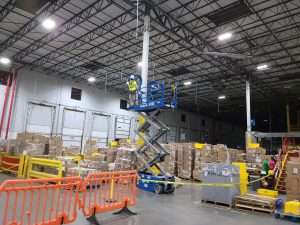 Electrician on a scissor lift performing overhead electrical work in a busy distribution warehouse